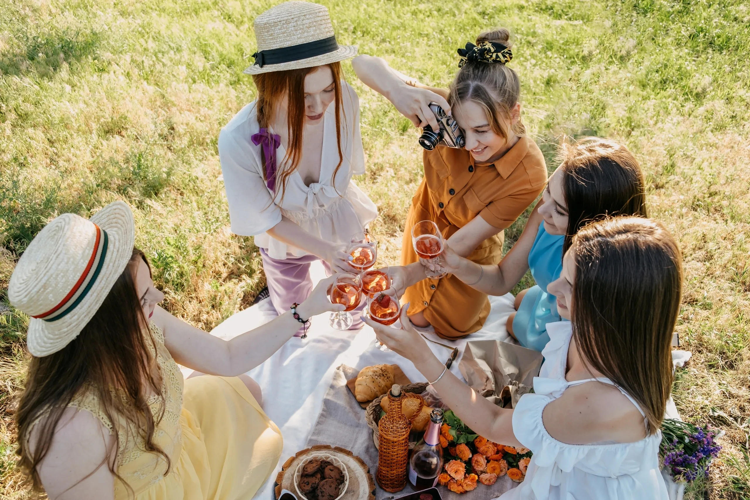 Five women enjoying a picnic, sitting on the grass and toasting with drinks, surrounded by an assortment of food.