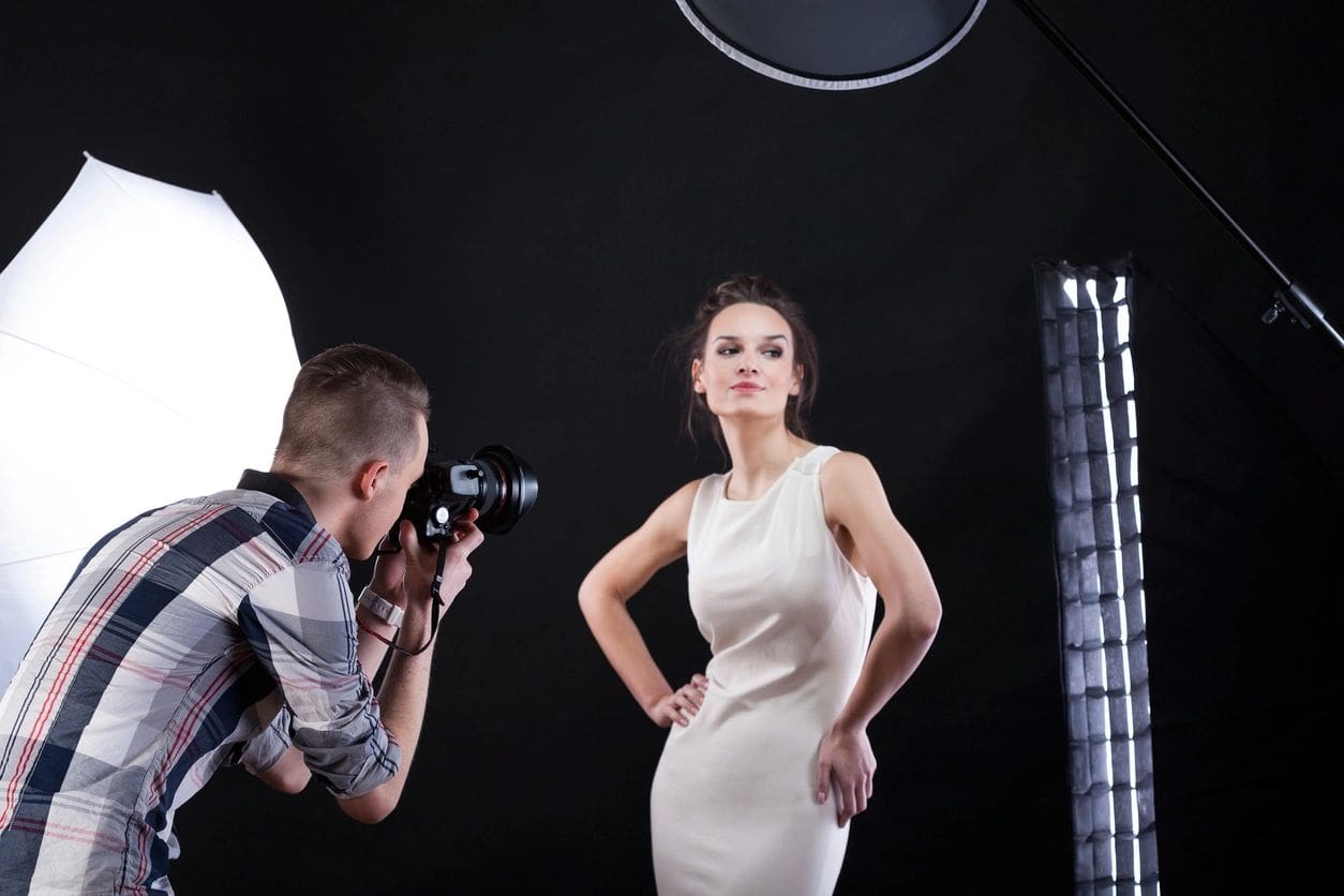 Photographer taking a picture of a female model in a white dress at a professional photo studio with various lighting equipment.